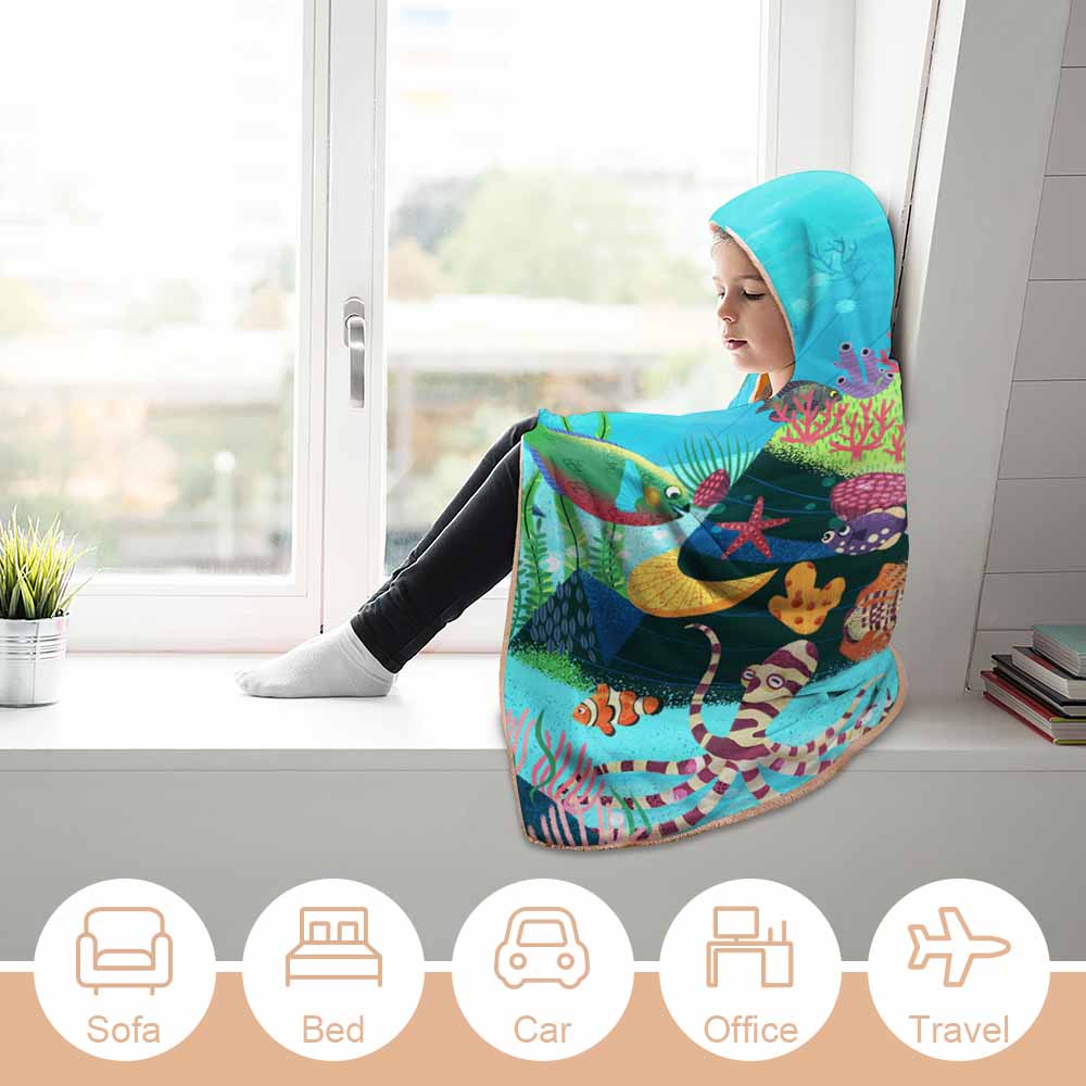 Cozy girl wearing an underwater scene cloak hooded blanket by a window, Arctic Velvet material.