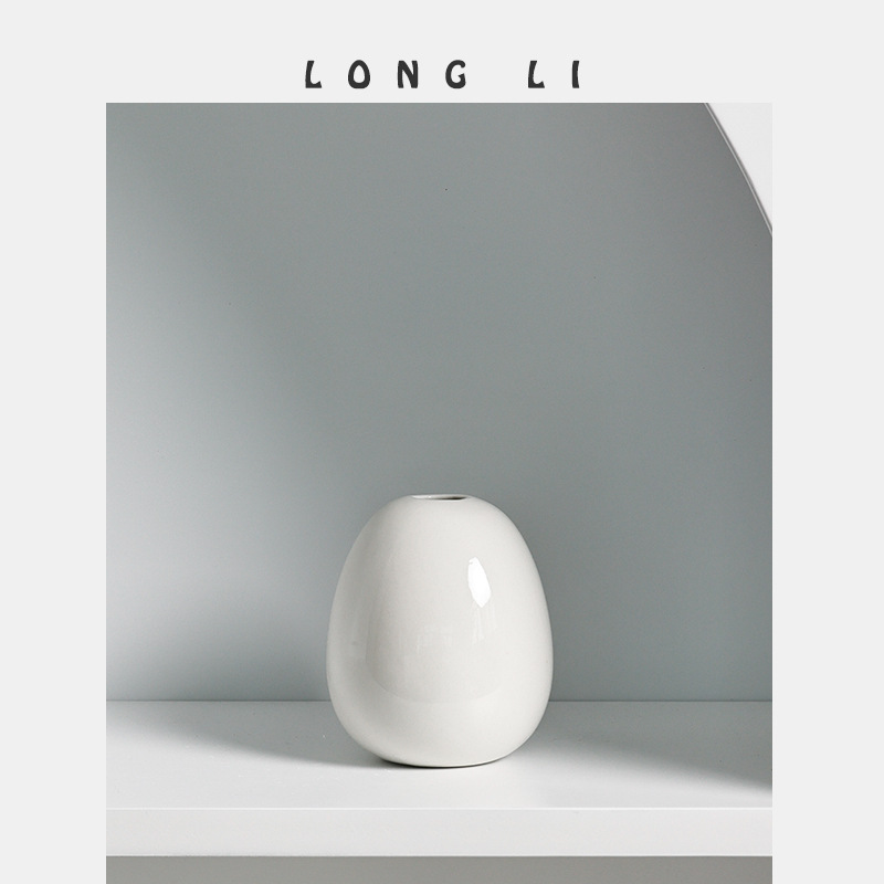 Egg-shaped white ceramic vase standing on a white shelf against a grey wall