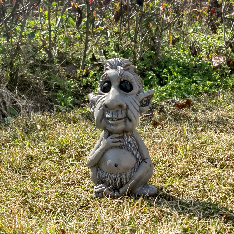 Small grey troll figurine standing in grass beside shrubs and leaves