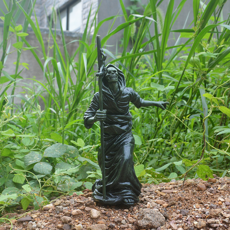 Black skeleton reaper guardian ornament holding a tall staff, standing among garden plants and gravel