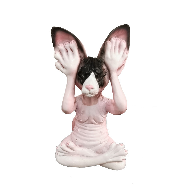 Pink and black-faced cat figurine with hands covering its ears on white