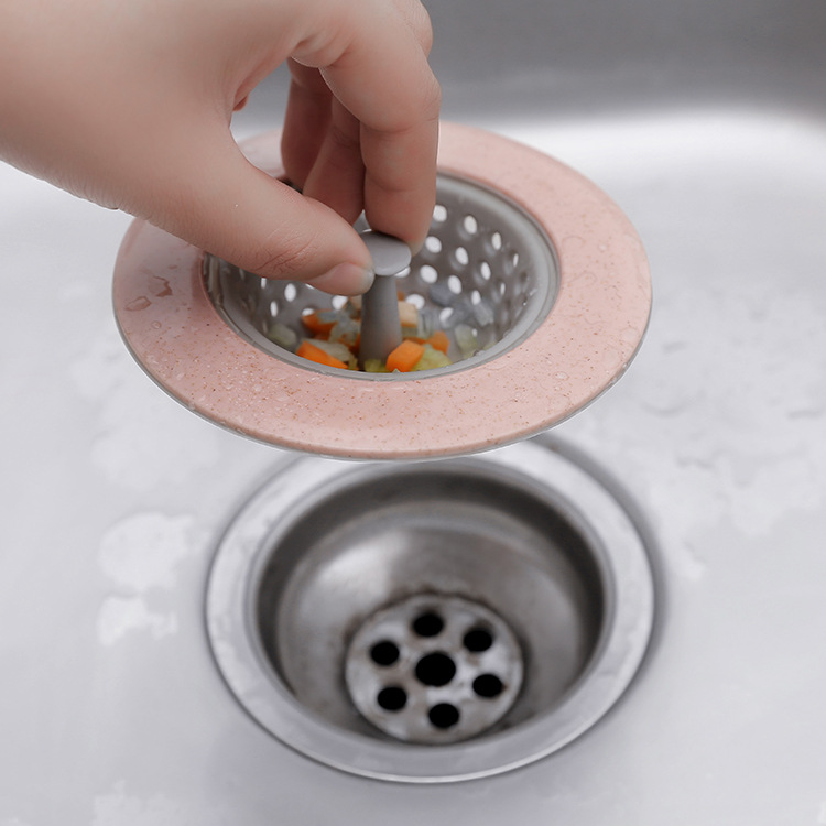 Hand lifting a pink sink strainer with trapped vegetable bits above a drain