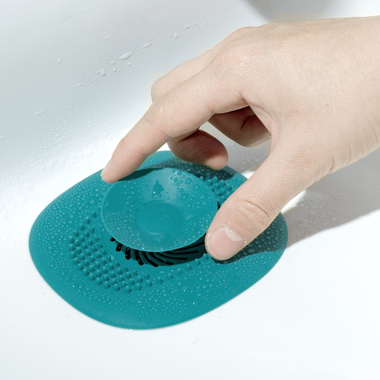 Hand pressing the teal drain cover's center plug into a wet white sink