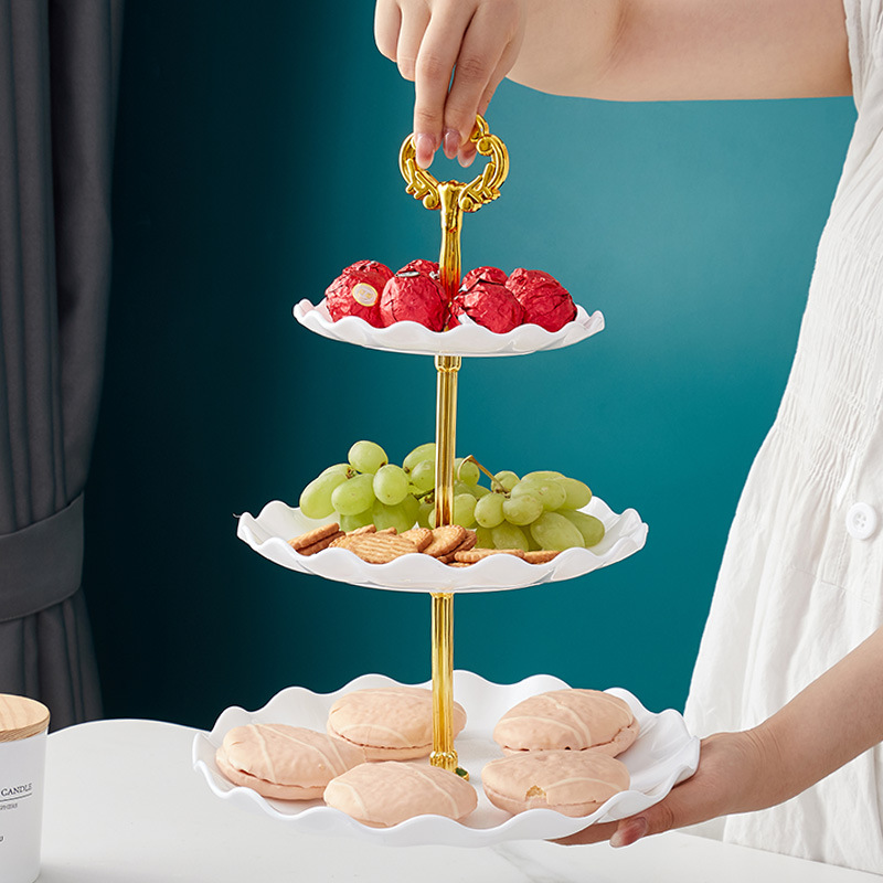 Person lifting a white three-tier stand by its gold handle, cookies on the bottom tier