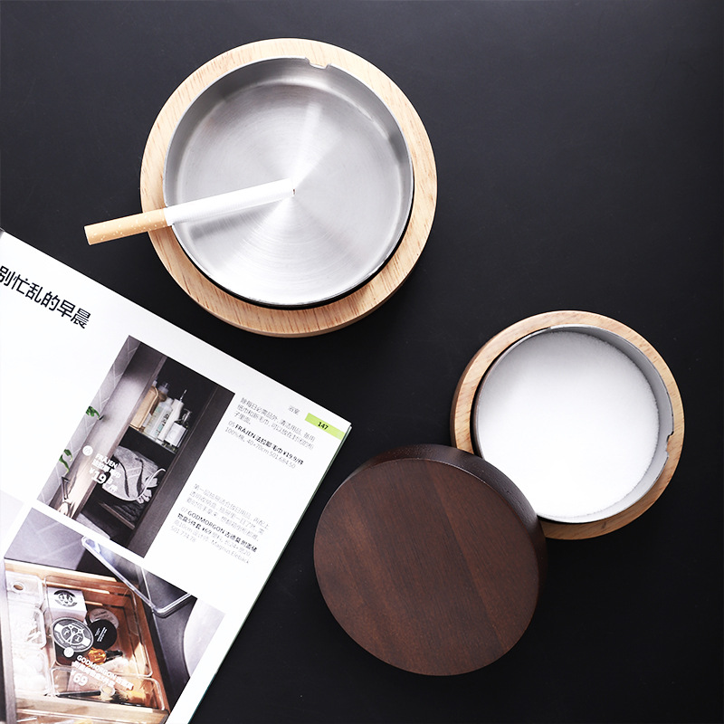 Open steel ashtray beside the dark wood lid and a bowl of white ash
