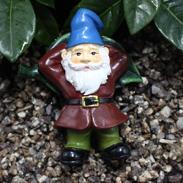 Blue-hat gnome with arms behind its head among green leaves on gravel