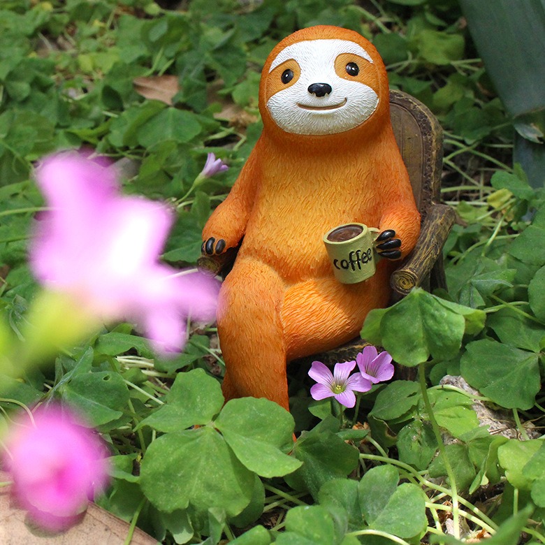 Sloth figurine with coffee mug set among garden plants in morning light