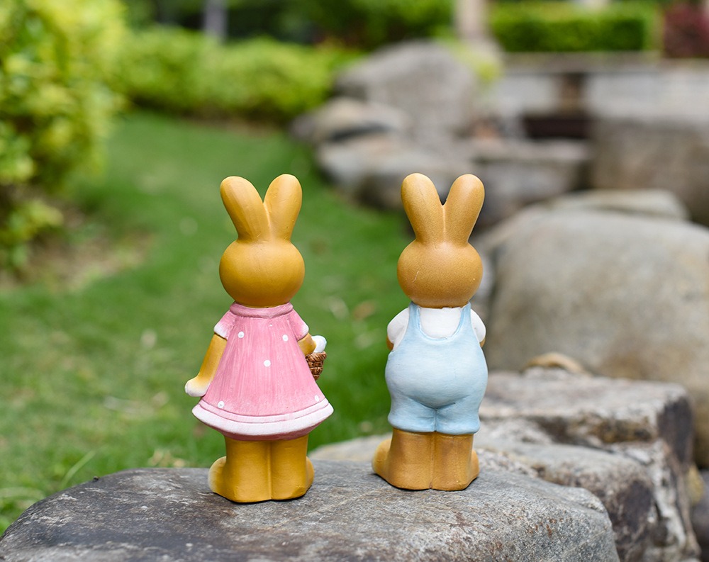 Back view of two ceramic bunnies sitting on a garden rock with shrubs