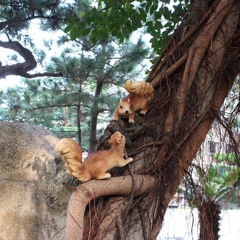 Two squirrel figurines climbing a tree trunk in a garden with pine trees behind