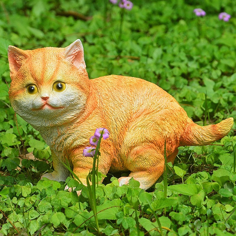 Orange tabby cat statue among green clover and purple flowers in a garden