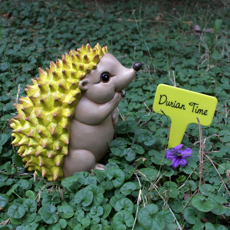 Hedgehog figurine in grass beside a yellow Durian Time plant stake
