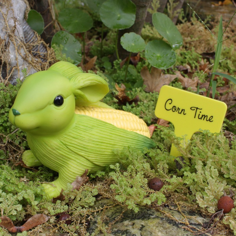 Corn rabbit ornament in a mossy garden bed beside a yellow stake