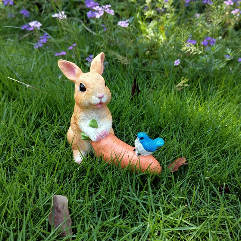 Tan rabbit with orange carrot sitting in a garden bed of grass and flowers