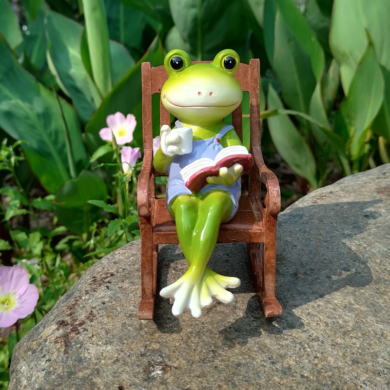 Frog figurine on a garden rock with pink flowers and leafy plants