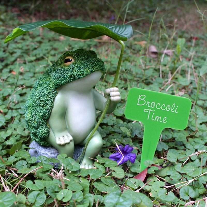Frog figurine in a garden bed beside the Broccoli Time plant stake