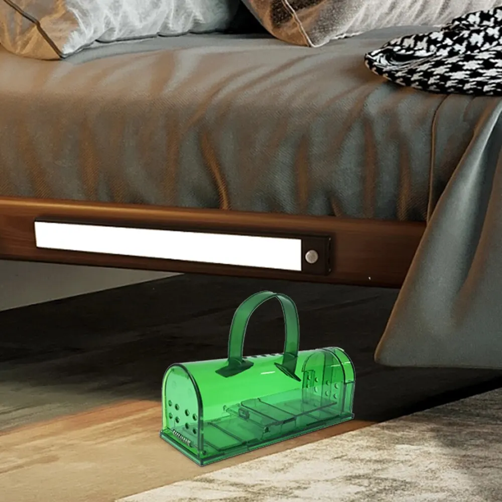 Green mousetrap set on bedroom floor under a bed frame