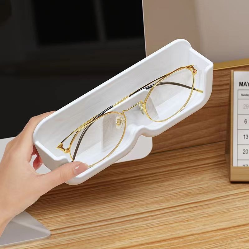 Hand holding a white glasses holder with gold-rimmed glasses beside a wooden desk