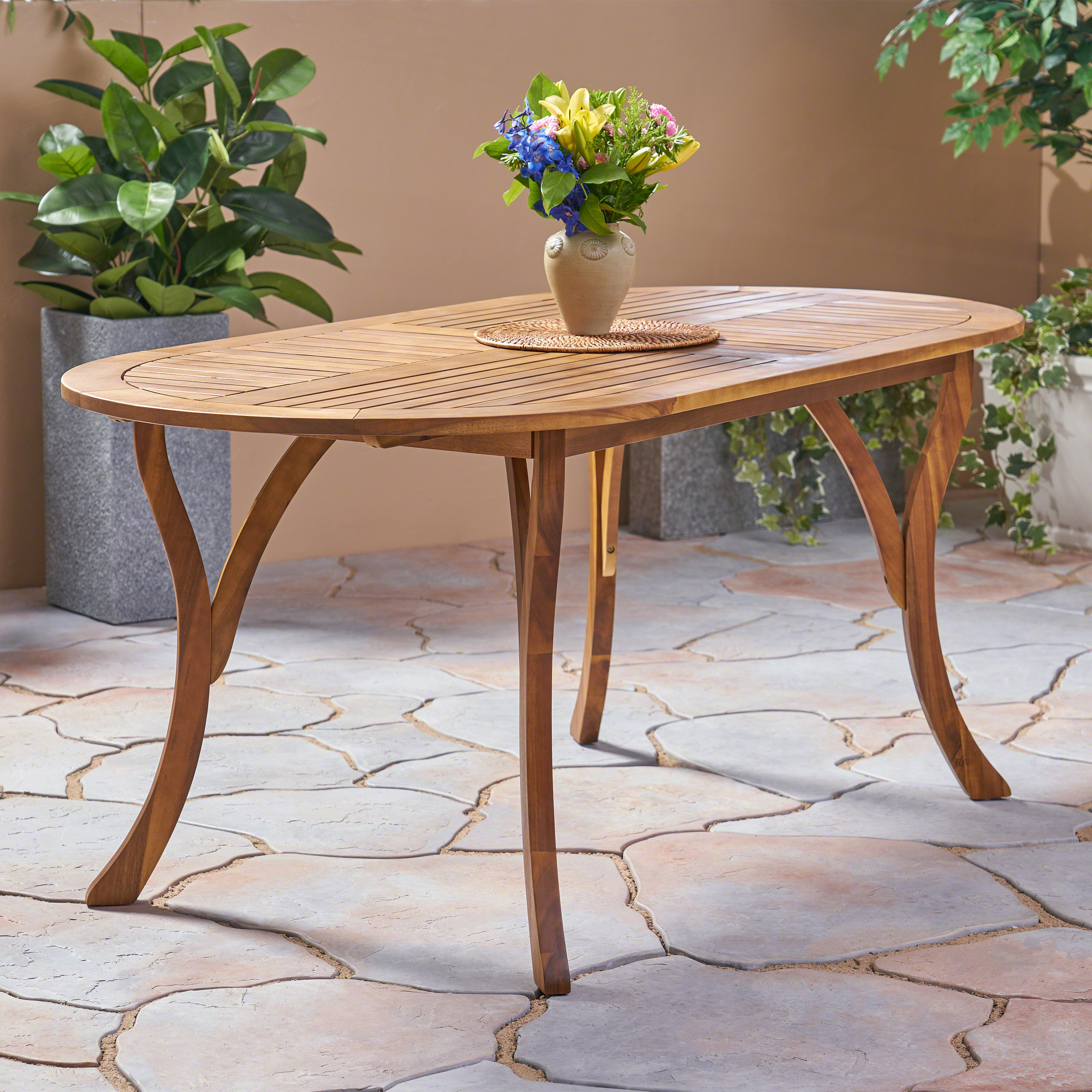 Oval acacia table set with a vase and woven placemat on a stone patio