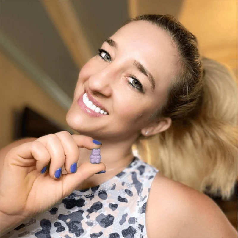 Woman with blue nail polish holding purple Sleep Deep gummies, smiling confidently in warm indoor light.