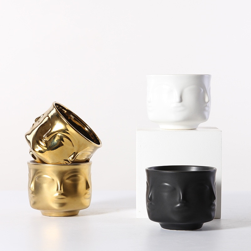 Stacked gold face planters beside a white and black face planter on a white cube